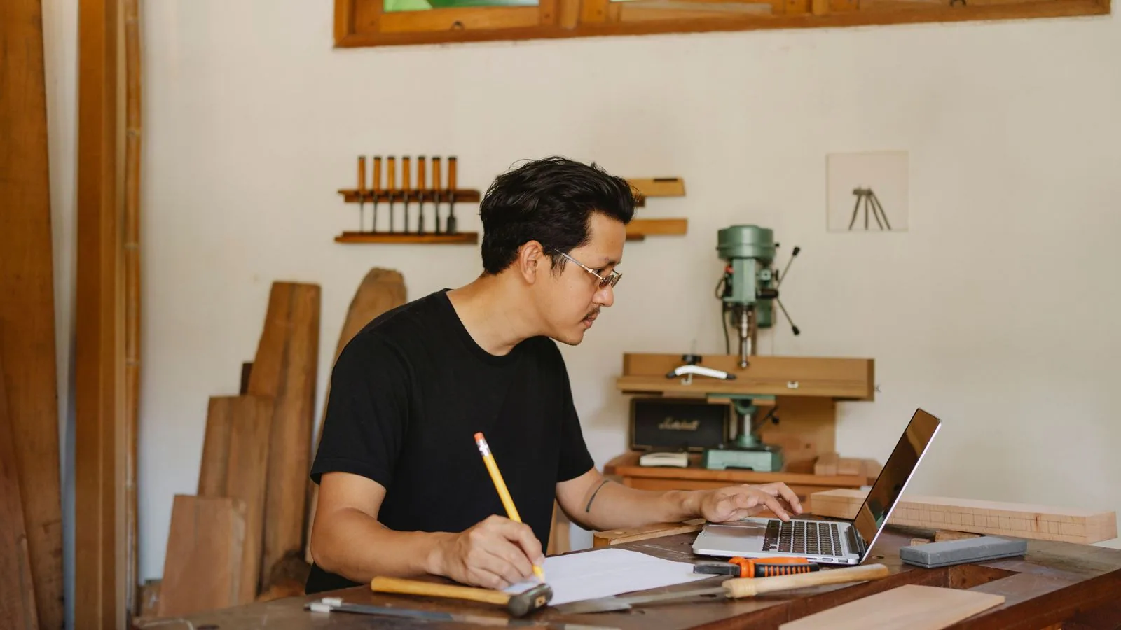 A woodworker using a laptop and notebook at a workshop bench