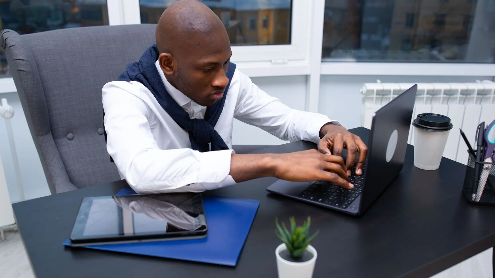 A man typing on a laptop beside a notebook